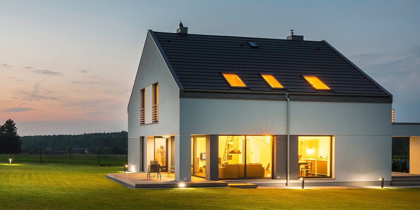 Ein modernes Haus mit großen Fenstern, das bei Dämmerung beleuchtet ist, umgeben von einem gepflegten Garten.